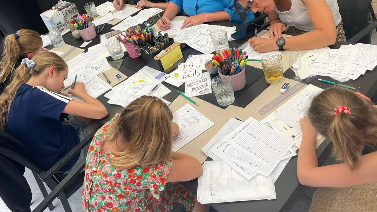 Deelnemers oefenen samen sier- en drukletters aan een tafel vol stiften en potloden