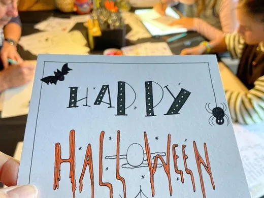 Een gehandletterd Halloween-kaartje met "Happy Halloween", omringd door deelnemers die aan een themaworkshop werken