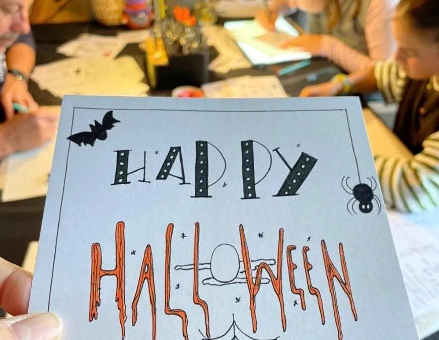 Een gehandletterd Halloween-kaartje met "Happy Halloween", omringd door deelnemers die aan een themaworkshop werken